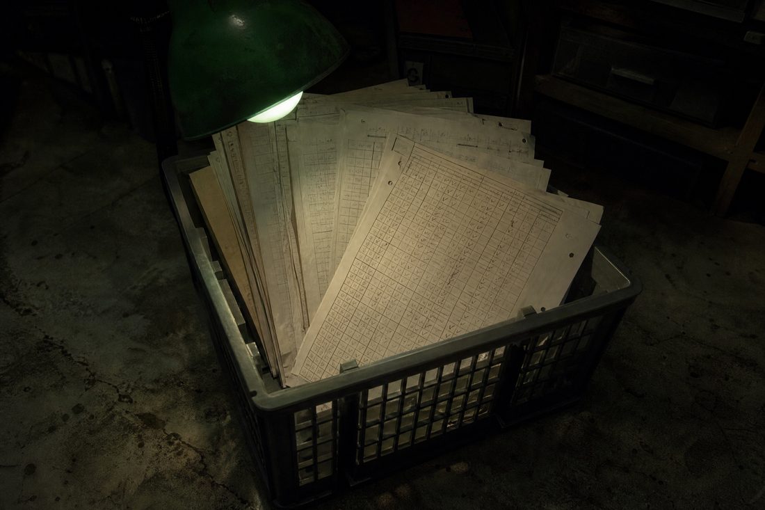 A plastic filing crate of loose printed pages dense with hand-drawn tables and pencil ticks, lit by a small green lamp on a concrete floor